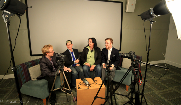 Eine Gruppe von drei Personen sitzt auf einem Sofa und gibt ein Interview, wobei eine Person links in einem schwarzen Mantel und Hose sitzt, eine Person in der Mitte in einem Mantel und blauer Krawatte ein Mikrofon hält und eine Person rechts strahlt. Ein Mikrofonrecorder befindet sich auf dem Tisch in der Mitte, Kameras sind auf der rechten und linken Seite und große Scheinwerfer sind vorhanden. Ein Projektionsschirm ist im Hintergrund zu sehen.