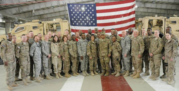 Eine Gruppe von Menschen, die mit einem Flaggenhintergrund, Fahrzeugen und Lichtern stehen und lächeln.