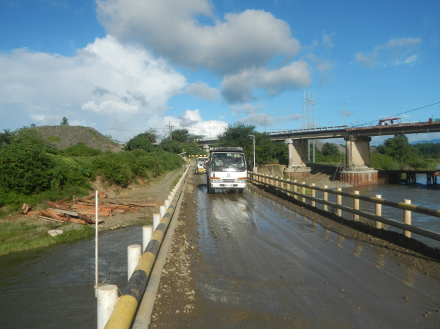Ein Laster fährt auf einer überfluteten Straße neben einer Brücke mit Geländern, mit Bäumen, Strommasten und einem bewölkten Himmel im Hintergrund.