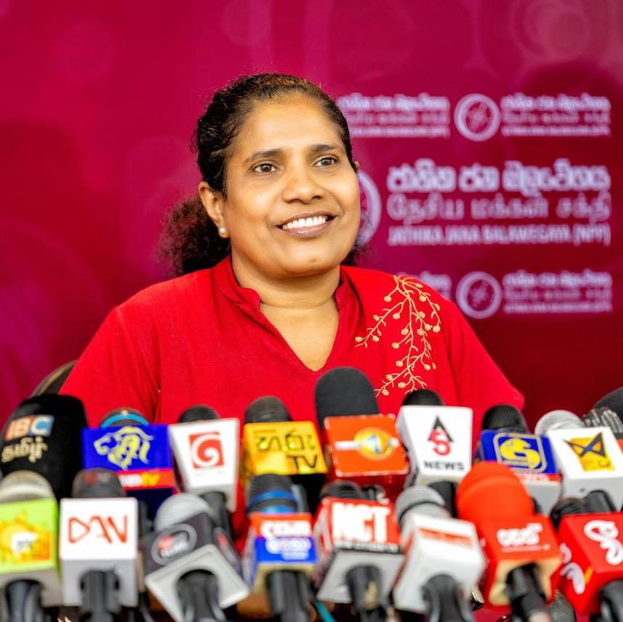 Eine Frau in einem roten Hemd sitzt an einem Tisch mit Mikrofonen, lächelt selbstbewusst, mit einem Banner dahinter, auf dem "Sri Lankas Gesundheitsminister" steht.