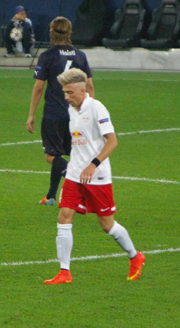 Ein Mann in einem weißen Hemd und roten Shorts spielt Fußball auf einem Feld, umgeben von Menschen, Stühlen und einer Wand, trägt Turnschuhe.