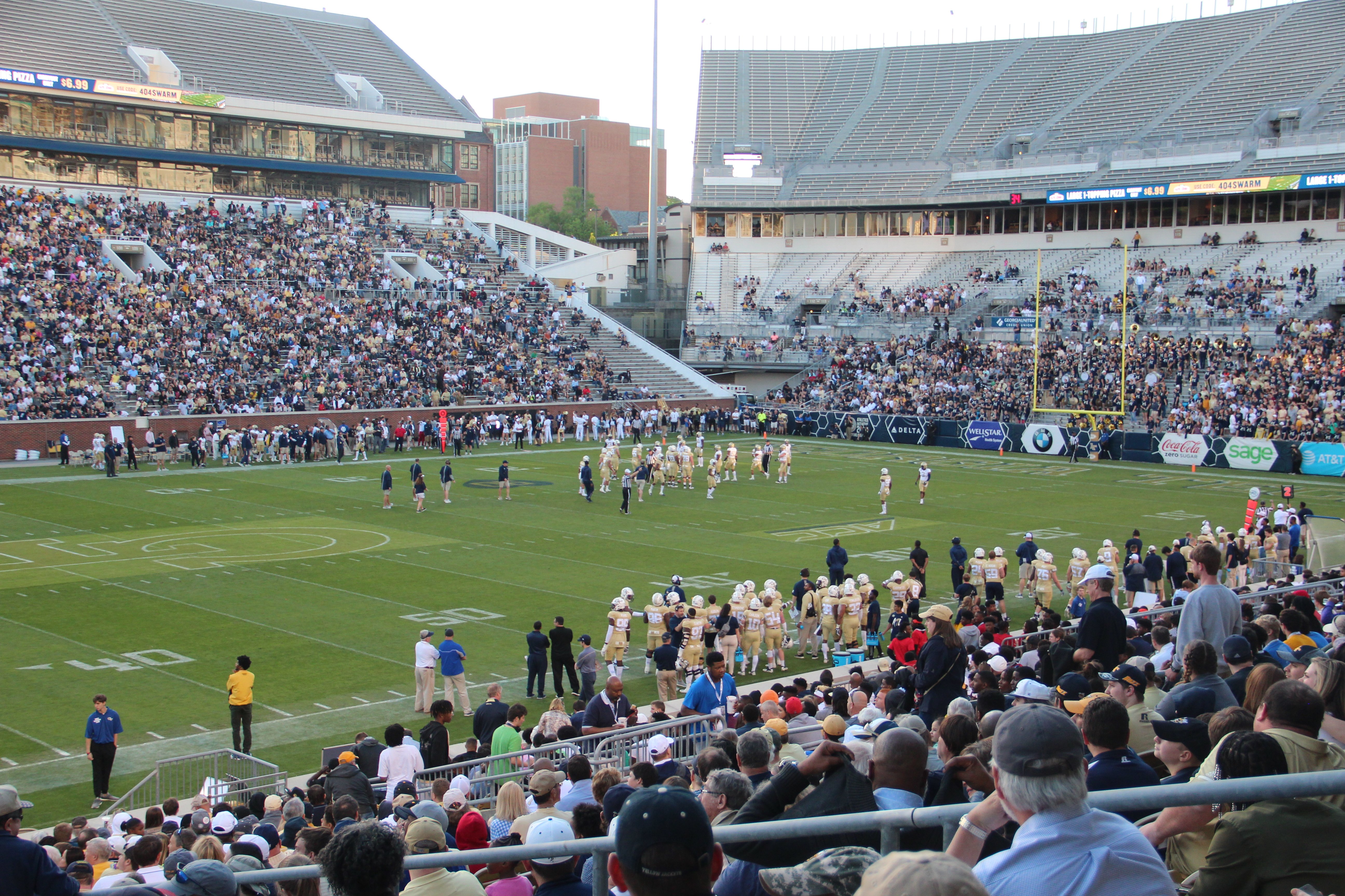 Ein Fußballspiel wird in einem Stadion mit Spielern auf dem Feld und Zuschauern in den Rängen unter einem klaren blauen Himmel gespielt.