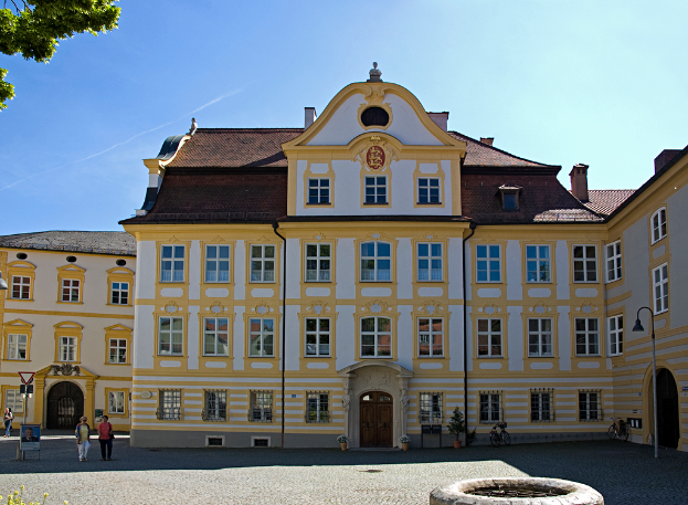 Ein weißes und gelbes Haus mit vielen Glasfenstern, mit zwei älteren Frauen, die auf einem Kopfsteinpflasterweg davor gehen.