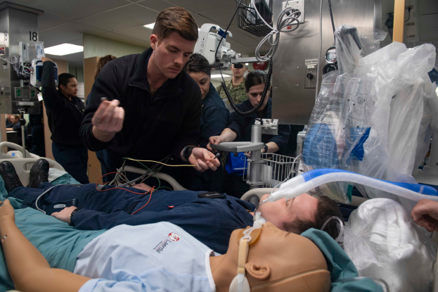 Ein Mann liegt auf einem Krankenhausbett neben einer Puppe, umgeben von einer Gruppe von Menschen und medizinischer Ausrüstung, in einer Trainingsübung innerhalb einer medizinischen Einrichtung.