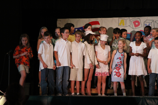 Mehrere Kinder stehen auf einer Bühne, zwei in der Mitte mit Mützen und ein Mikrofonständer links daneben, mit einem Banner im Hintergrund.