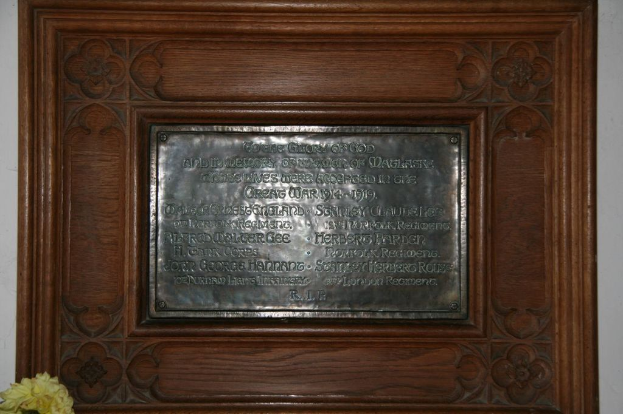 Eine Gedenktafel mit Text ist an einer braunen Wand oder einer Holztür angebracht, mit einem gelben Objekt, das einer Vase ähnelt, in der linken unteren Ecke.