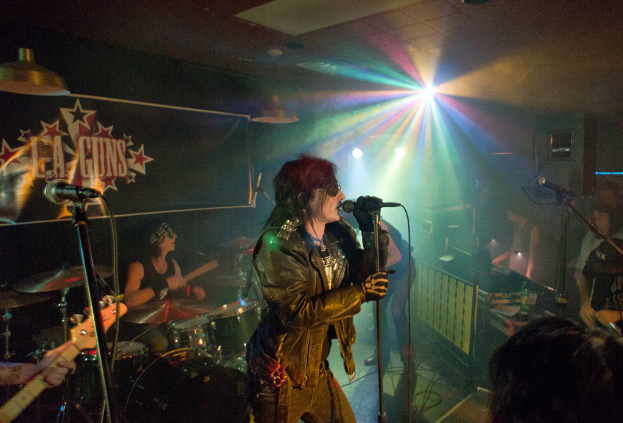 Eine Frau in einer schwarzen Jacke singt in ein Mikrofon auf der Bühne, mit einem Mann in Sonnenbrille und einem Headband, der hinter ihr Bassgitarre spielt, unter einem Schild, auf dem "LA Guns" steht und bunten Scheinwerfern.