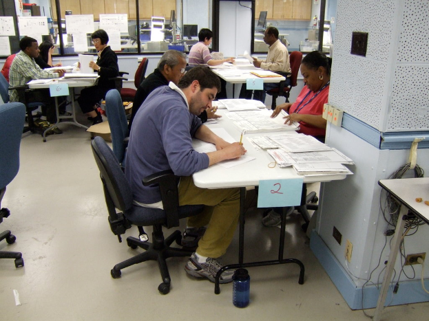 Mitarbeiter an Schreibtischen mit Papieren, einige schreiben und andere erklären, mit Glasfenstern und Monitoren im Hintergrund.