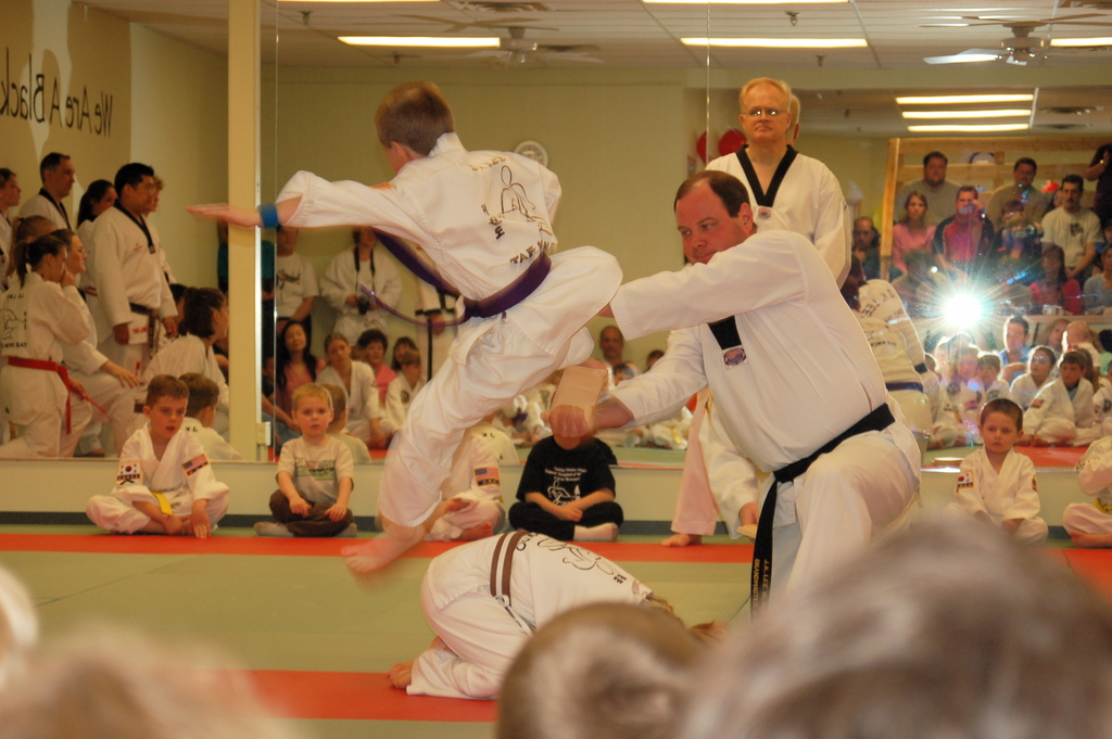 Zwei Personen kämpfen in einem Raum mit Kindern auf dem Boden, während im Hintergrund eine Gruppe von Menschen, eine Kamera, eine Uhr, eine Wand, ein Dach, Deckenlampen, ein Ventilator, eine Säule und eine weitere Wand zu sehen sind.