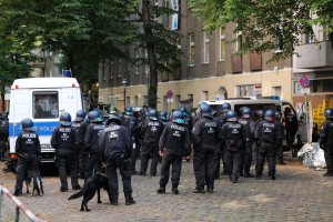 Eine Gruppe von Polizisten in schwarzen Uniformen und Helmen steht vor einer Menge, mit einem schwarzen Hund im Vordergrund; im Hintergrund gibt es Fahrzeuge, Bäume, Gebäude und ein Geländer.