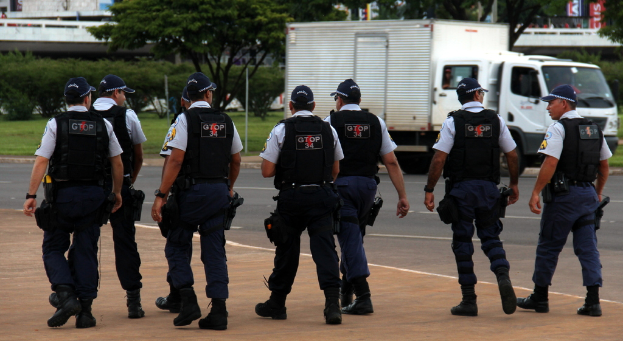 Eine Gruppe von Offizieren in Uniform, einschließlich Mützen, Hemden, kugelsicheren Westen, Uhren, Hosen und Schuhen, die auf einer Straße mit einem geparkten Lastwagen auf der rechten Seite neben einem Zaun gehen. Im Hintergrund sind Gebäude, Bäume, Pflanzen, Pfosten und Banner zu sehen, mit einer Brücke in der oberen rechten Ecke.