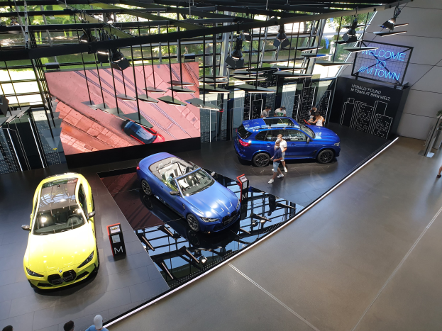 Eine Gruppe von Menschen, die im BMW Museum in München stehen, mit ausgestelltem Auto auf der linken Seite und einem Schild mit der Aufschrift "BMW Museum München" auf der rechten Seite, mit Blick auf eine außerstliche Stadtlandschaft durch Glasfenster.