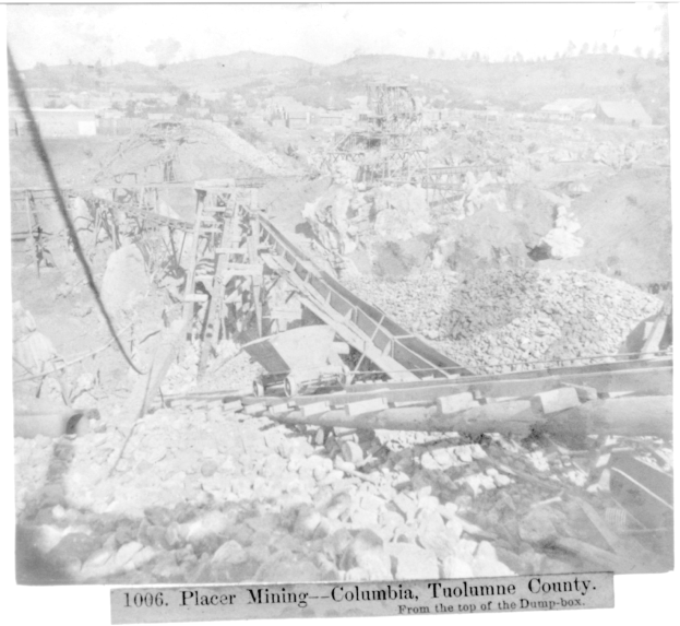 Ein Schwarz-Weiß-Foto eines Kohlebergwerks in Kolumbien, das ein paar Gebäude, Bäume und Berge im Hintergrund zeigt, mit Text am unteren Rand des Bildes.