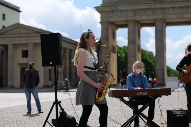 Eine Frau spielt Saxofon vor dem Brandenburger Tor in Berlin, Deutschland, mit einer Person, die ein Piano spielt, einer anderen, die eine Gitarre hält, und einem Lautsprecher im Vordergrund, während Gebäude, Bäume und Wolken im Hintergrund zu sehen sind.
