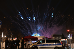 Ein Polizeiwagen parkt vor einer Menschenmenge, umgeben von Fahrrädern, Zelten, Laternenpfählen, Schildern, Verkehrszeichen, Bäumen, Gebäuden und einer Statue im Hintergrund, unter einem von Feuerwerk erhellten Himmel.