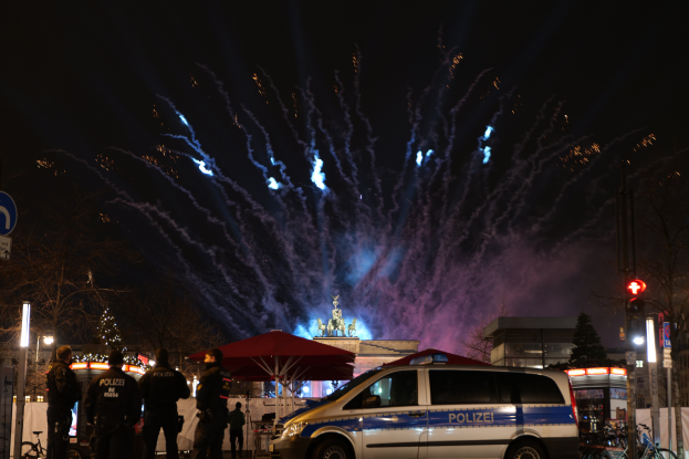 Ein Polizeiwagen parkt vor einer Menschenmenge, umgeben von Fahrrädern, Zelten, Laternenpfählen, Schildern, Verkehrszeichen, Bäumen, Gebäuden und einer Statue im Hintergrund, unter einem von Feuerwerk erhellten Himmel.