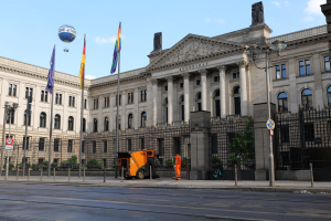 Großes modernes Regierungsgebäude mit vielen Fenstern, umgeben von Straßenmöbeln und Fahrzeugen, mit Menschen auf dem Bürgersteig und wehenden Flaggen davor.