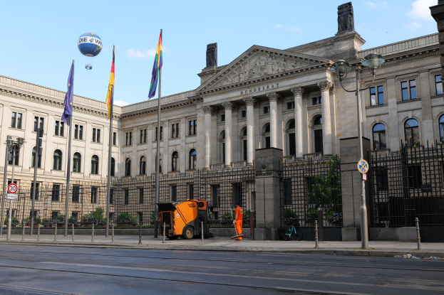Großes modernes Regierungsgebäude mit vielen Fenstern, umgeben von Straßenmöbeln und Fahrzeugen, mit Menschen auf dem Bürgersteig und wehenden Flaggen davor.