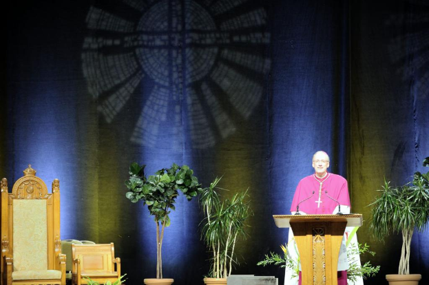 Ein Mann in einem pinken und weißen Kleid steht neben einem Rednerpult in einem Auditorium, mit Pflanzen und zwei Stühlen daneben und einer bunten Wand im Hintergrund.