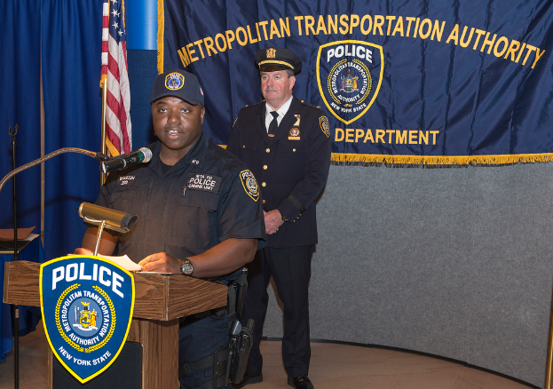 Zwei Polizeibeamte stehen vor einem Rednerpult, einer hält Papers und spricht in ein Mikrofon, mit einer Fahne, einem Tisch und einer Tafel mit Symbolen und Text im Hintergrund.