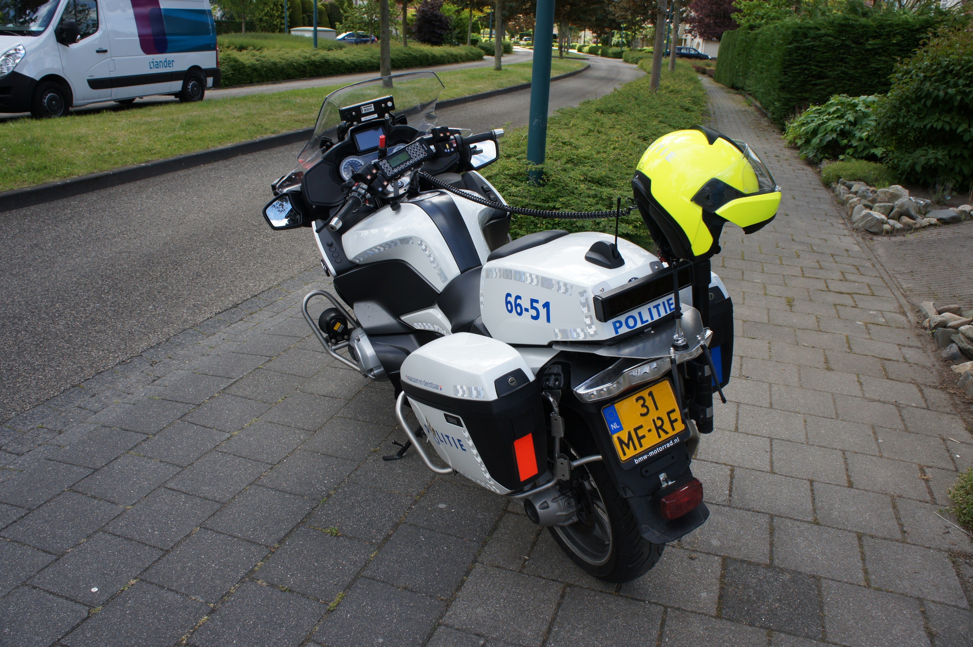 Eine Polizei-Motorrad am Straßenrand mit einem Helm darauf, umgeben von Bäumen, Pfählen, Gebäuden, Pflanzen, Gras, Steinen und anderen Fahrzeugen im Hintergrund.