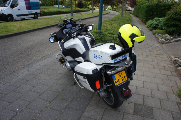 Eine Polizei-Motorrad am Straßenrand mit einem Helm darauf, umgeben von Bäumen, Pfählen, Gebäuden, Pflanzen, Gras, Steinen und anderen Fahrzeugen im Hintergrund.
