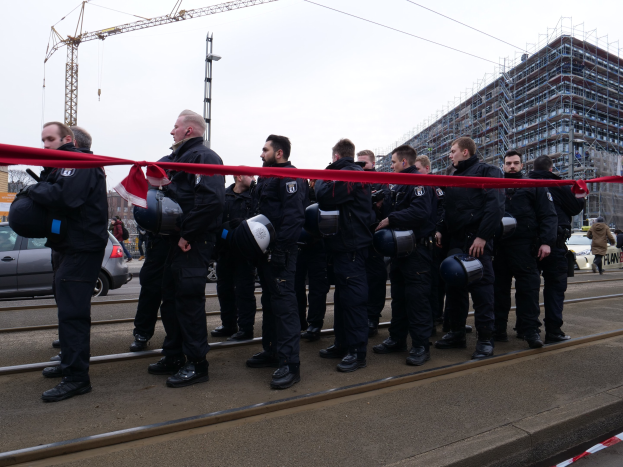 Eine Gruppe von Polizisten in schwarzen Uniformen und Helmen steht in einer Reihe auf einem Bahngleis mit einem roten Band vor ihnen, während Fahrzeuge, Fußgänger, Gebäude und Strommasten im Hintergrund unter einem klaren blauen Himmel zu sehen sind.