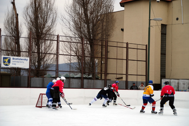 Menschen, die Eis Hockey auf einem Eisplatz mit Gebäuden, Bäumen, einer Straßenlaterne, einem Namensschild und Zäunen im Hintergrund unter einem Himmel spielen.