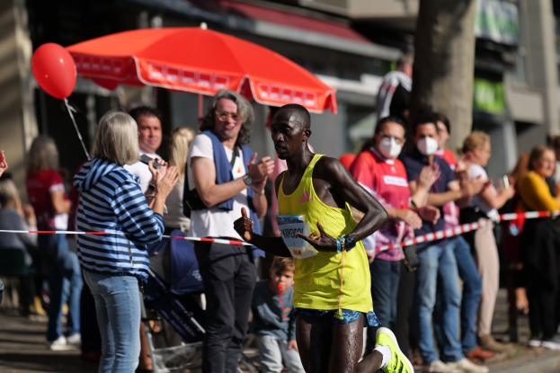 Ein Mann in einem gelben T-Shirt läuft bei einem Marathon, umgeben von einer jubelnden Menge, mit einem Schirm und einem Kinderwagen im Hintergrund.