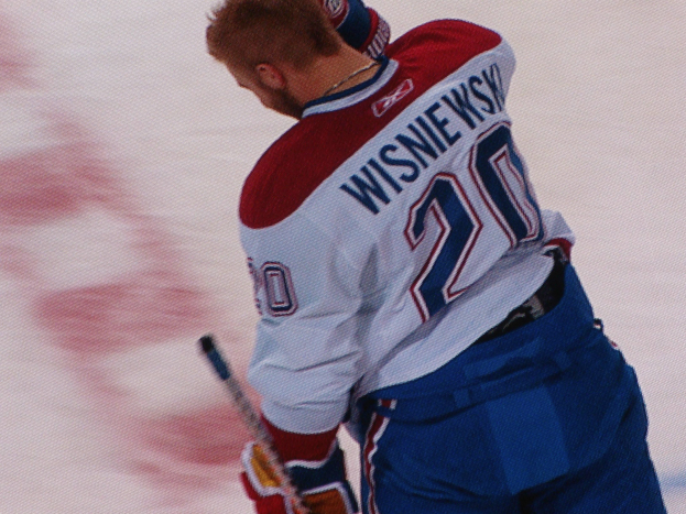 Ein Eishockeyspieler in einem "Wisniewski"-Trikot steht auf dem Eis und hält einen Eishockeyschläger mit konzentrierter und entschlossener Miene.