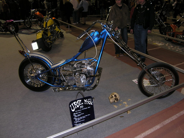 Mehrere Motorräder und ein Schädel sind innerhalb eines eingezäunten Bereichs zu sehen, mit einem Plakat und einer Tafel mit Text; einige Menschen stehen hinter dem Zaun.