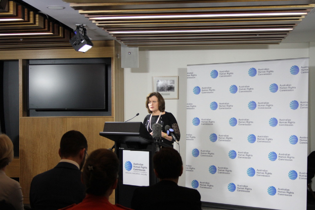 Eine Frau steht auf einer Bühne und spricht in ein Mikrofon, vor ihr sitzen Menschen auf Stühlen, an der Wand hängt ein Banner, auf dem Holzwand steht ein Fernseher und an der Decke leuchten Lampen.