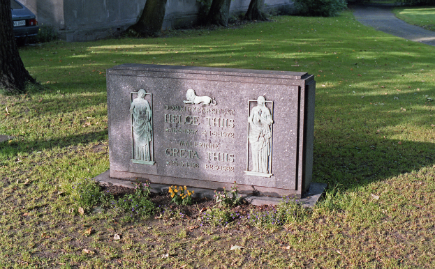 Ein Gedenkstein mit Bildern und Text, umgeben von Gras, einer Straße, Bäumen, einem geparkten Auto und einer Wand im Hintergrund.