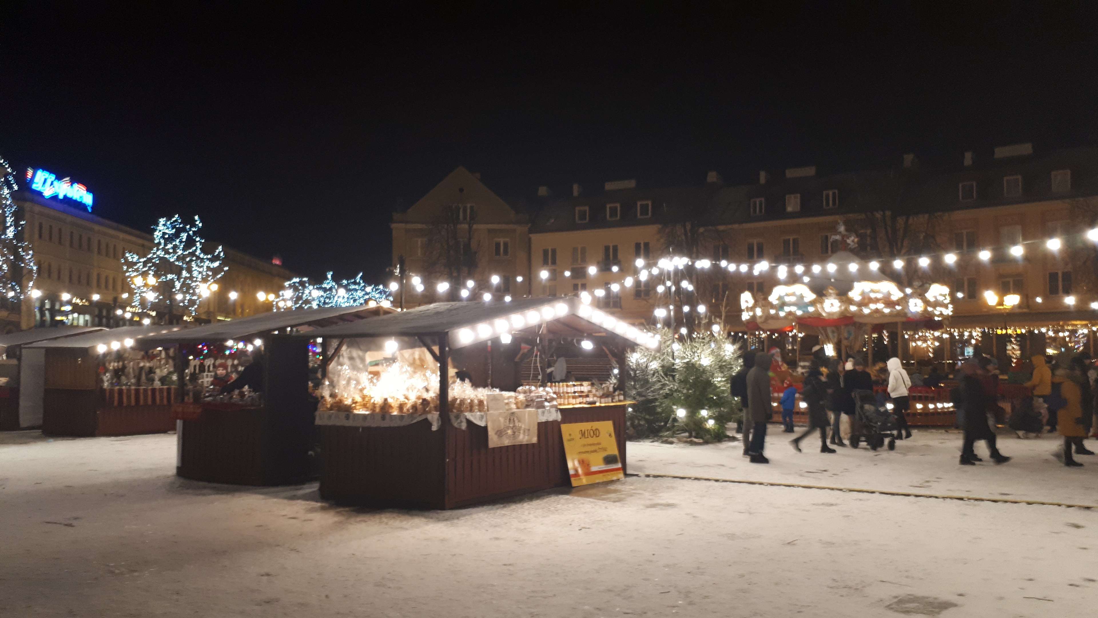 Weihnachtsmarkt-Szene bei Nacht mit Menschen, Buden, Pflanzen, Bäumen, Gebäuden und Schildern in einer verschneiten Umgebung.