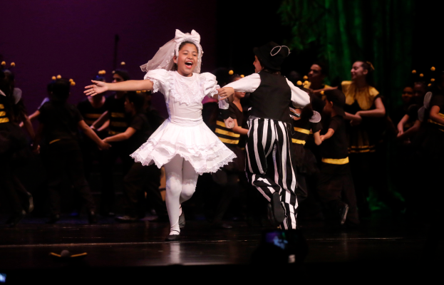 Ein junges Mädchen in einem weißen Kleid und ein Mann in einem schwarz-weißen gestreiften Anzug tanzen auf einer Bühne, umgeben von kostümierten Menschen, mit einem grünen Vorhang und einer violetten Wand im Hintergrund.