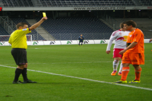 Ein Schiedsrichter zeigt einem Fußballspieler auf dem Feld eine gelbe Karte, umgeben von einer Gruppe von Menschen und einem Torpfosten, einem Zaun, einem Schild, Metallrahmen, einem Bildschirm, Stühlen und einer Decke mit Deckenleuchten im Hintergrund.