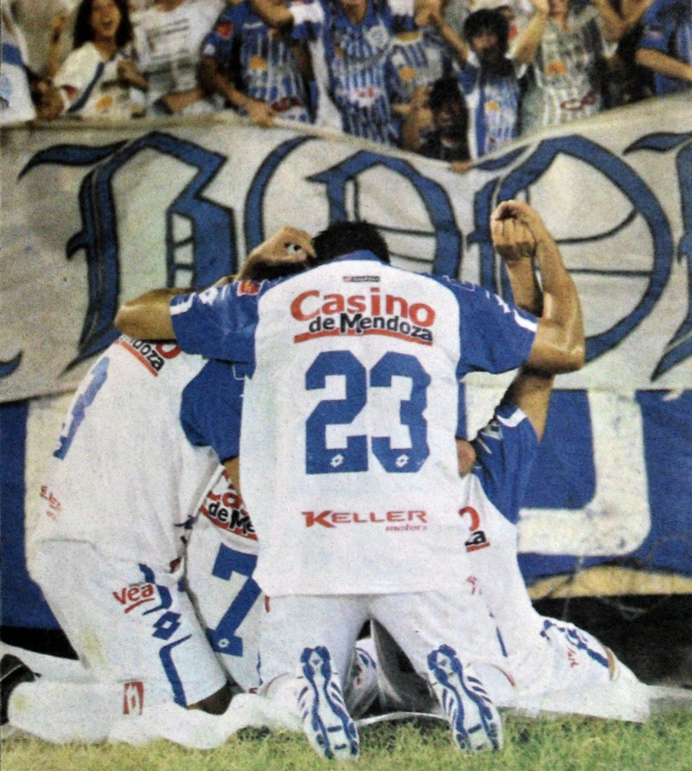 Eine Gruppe von Fussballspielern feiert ein Tor auf dem Boden vor einer jubelnden Menge, mit einem teilweise sichtbaren Banner im Hintergrund.