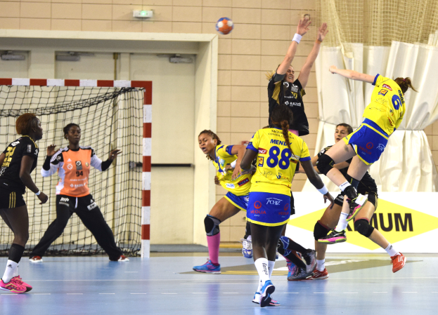 Eine Gruppe von Frauen beim Handballspielen auf einem Feld, mit einem Ball in der Luft, einem Torpfosten mit Netz, einer Fahne mit Text, einem Vorhang und einer Wand im Hintergrund.
