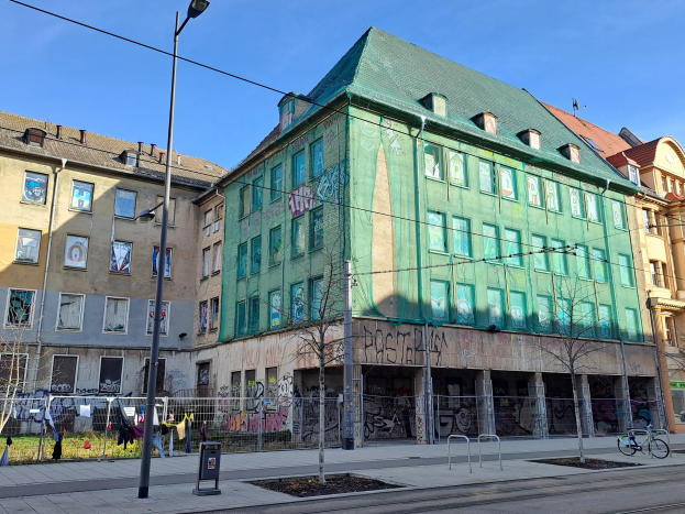 Großes grünes Gebäude mit Graffiti an der Seite, Teil des Berlin Wall Projects in Berlin, Deutschland, umgeben von städtischen Elementen wie Straßenlaternen, Fahrzeugen und Fußgängern.