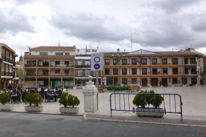 Ein belebter Stadtplatz mit Menschen, die sitzen und stehen, Topfpflanzen, Metallabsperrungen, ein Schild an einem Pfahl, Straßenlaternen mit Fahnen, Gebäude und eine bewölkte Himmel.