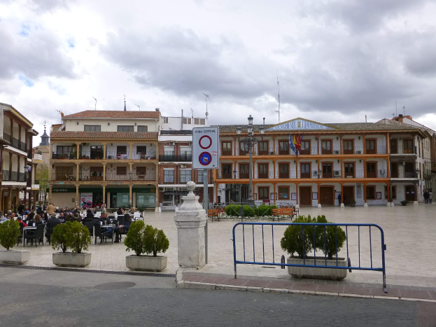 Ein belebter Stadtplatz mit Menschen, die sitzen und stehen, Topfpflanzen, Metallabsperrungen, ein Schild an einem Pfahl, Straßenlaternen mit Fahnen, Gebäude und eine bewölkte Himmel.