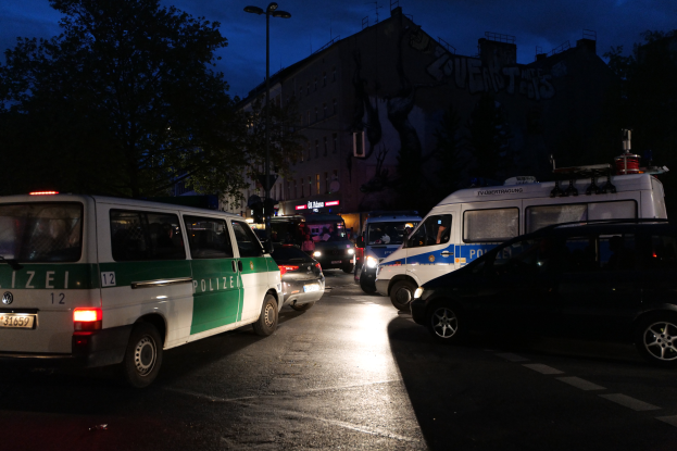 Eine Gruppe von Polizeiwagen, einschließlich eines Vans, Autos und Rades, die nachts an der Seite einer Straße geparkt sind mit Gebäuden, Bäumen, Laternenmasten und einem sternenklaren Himmel im Hintergrund.