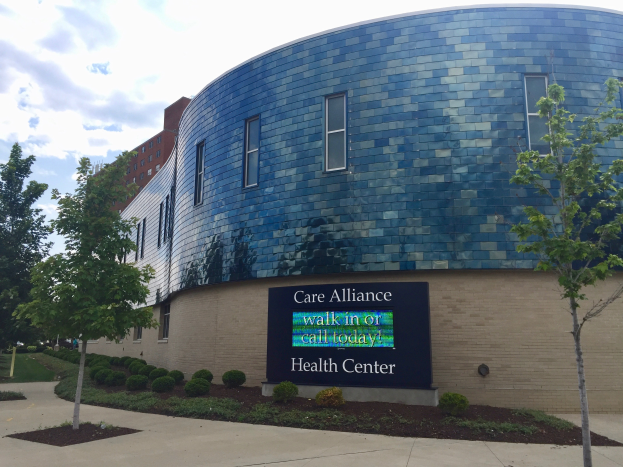 Ein Gebäude mit Fenstern und einem Schild, umgeben von Bäumen, Pflanzen und Gras, mit einem Weg, der darauf zuführt, unter einem bewölkten Himmel.