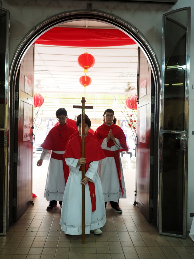 Menschen, die eine Kreuzes tragen, während sie durch eine Kathedrale gehen, mit Türen links und rechts und Blumen und dekorativen Elementen im Hintergrund.