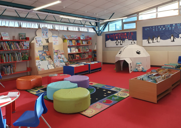Eine hell erleuchtete Kinderbibliothek mit Bücherregalen, Tischen, Stühlen, Teppichen, Wandtafeln und Deckenlampen.
