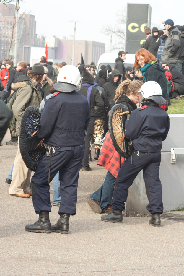 Eine Gruppe von Menschen, die auf einer Straße gehen, mit zwei Personen in der Mitte, die Polizeibeamte ähneln, Gebäuden im Hintergrund und Boden unten.