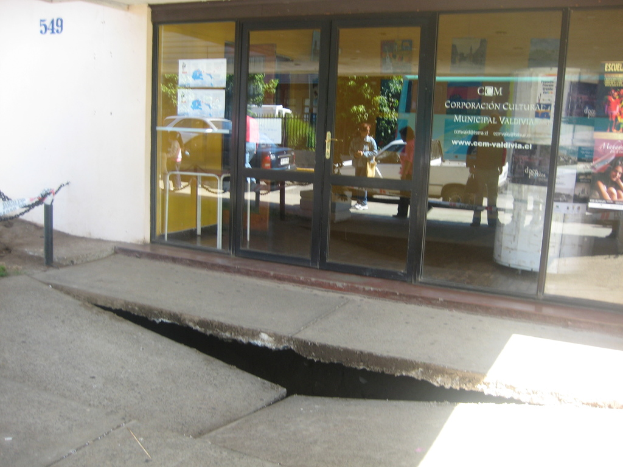 Eine gebrochene Oberfläche im Vordergrund mit einer Glastür, Wand und Pfahl im Hintergrund, die sich bewegende Fahrzeuge und einige stehende Personen auf der Straße spiegeln.