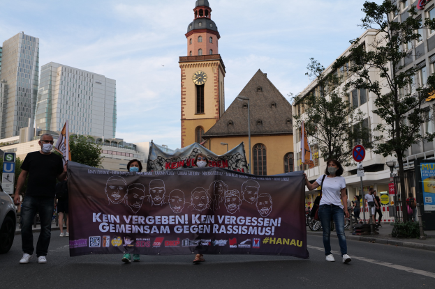 Eine Gruppe von Menschen marschiert auf einer Straße und hält ein Banner hoch, mit einem geparkten Auto auf der linken Seite, Gebäuden und Bäumen im Hintergrund und einem klaren blauen Himmel darüber.