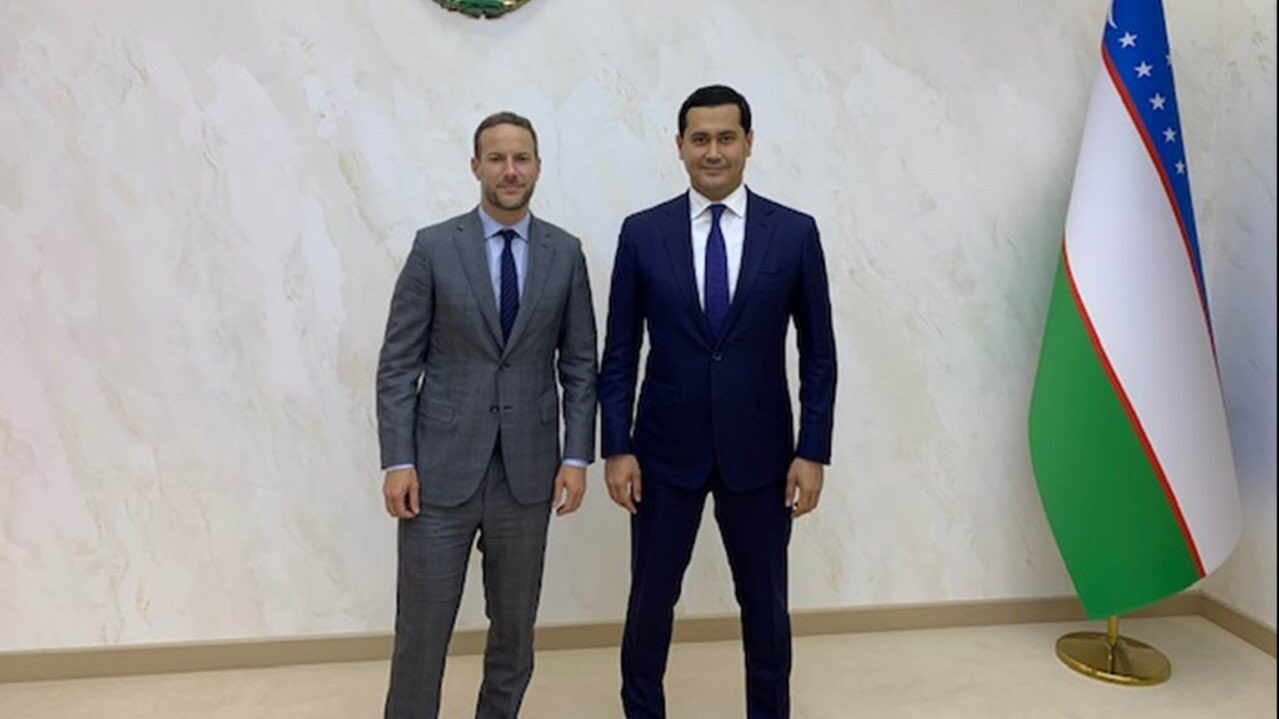 Bulgariens Premierminister und Außenminister, beide im Anzug, stehen lachend nebeneinander vor einer Flagge, mit einem Flaggenständer rechts daneben und einem Objekt an der Wand im Hintergrund.