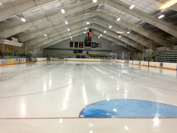 Große Indoor-Eisfläche mit Deckenbeleuchtung, Texttafeln, einer Wand, Treppen mit Geländern und einigen Menschen, was auf eine Sportanlage hinweist, die likely für Hockey verwendet wird.